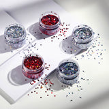 Five small jars of glitter on a white surface with scattered glitter.